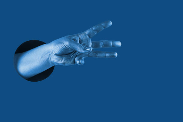 istock Hand shows  different gestures and symbols from the hole on a blue paper background. Template for design. Trend color of 2020 concept. 1205291504
