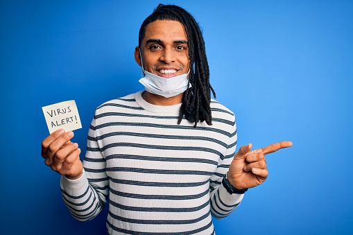 African american man with dreadlocks wearing medical mask holding virus alert reminder very happy pointing with hand and finger to the side