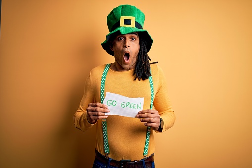 Young african american man wearing green hat holding banner celebrating saint patricks day scared in shock with a surprise face, afraid and excited with fear expression