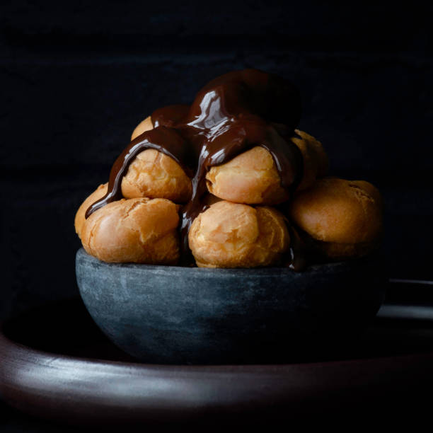 A tempting assortment of golden cream puffs topped with rich, glossy chocolate glaze, beautifully arranged in individual wrappers. These decadent pastries are perfect for dessert tables, celebrations, or as a delightful treat for chocolate lovers. The close-up view showcases their fluffy texture and indulgent toppings, creating a feast for the eyes and the palate