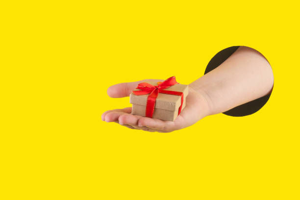 Hand holds a gift box made of natural materials through a hole in a yellow paper background. stock photo
