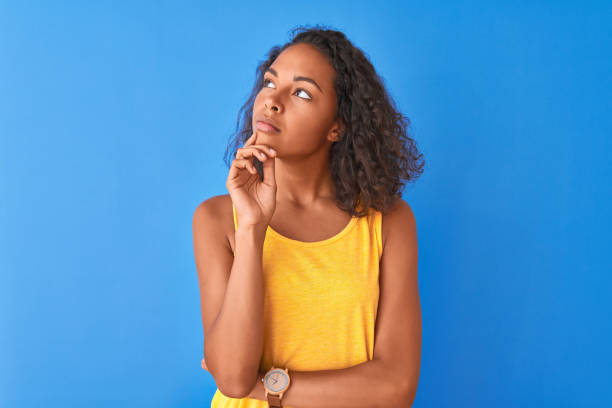 jovem brasileira vestindo camiseta amarela em pé sobre fundo azul isolado com a mão no queixo pensando em pergunta, expressão pensativa. sorrindo com o rosto atencioso. conceito de dúvida. - teenage girls ideas blue yellow - fotografias e filmes do acervo