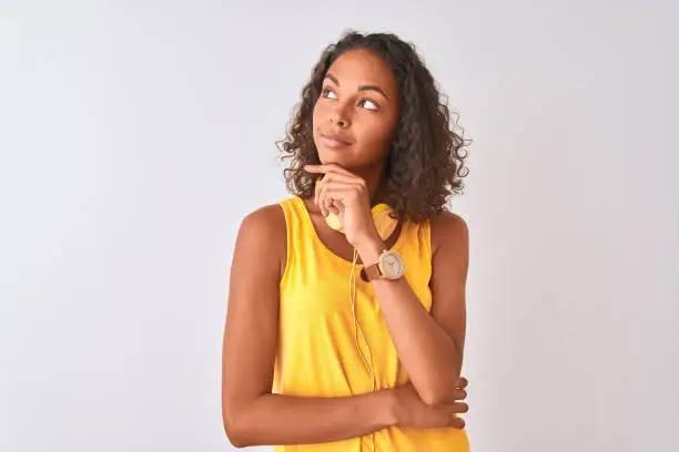 Young brazilian woman wearing yellow headphones over isolated white background with hand on chin thinking about question, pensive expression. Smiling with thoughtful face. Doubt concept.