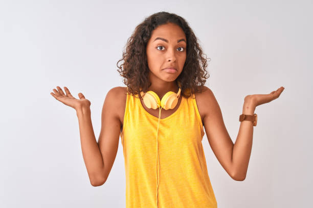 young brazilian woman wearing yellow headphones over isolated white background clueless and confused expression with arms and hands raised. doubt concept. - head and shoulders audio imagens e fotografias de stock