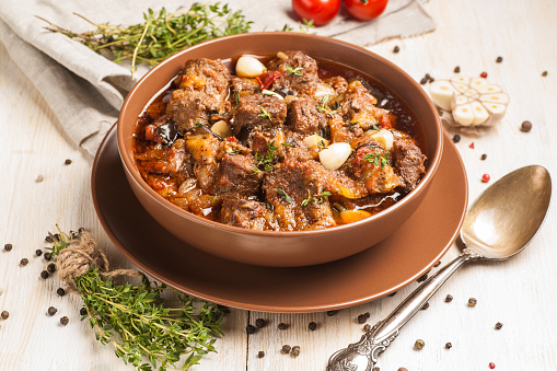 Braised meat with vegetables in a thick sauce. A large portion in a deep plate is ready to eat. Rustic decoration on a light wooden background. Close up.