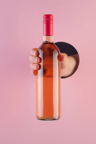 Hand holds a bottle of rose wine through a hole in paper pink background. stock photo