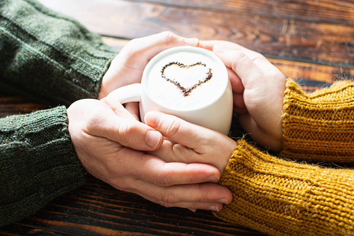 Close up of a couple holding hands on a mug of coffee with a cocoa heart on milk foam