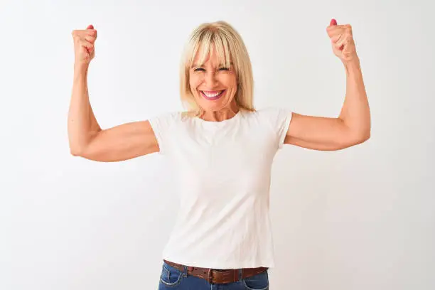 Middle age woman wearing casual t-shirt standing over isolated white background showing arms muscles smiling proud. Fitness concept.