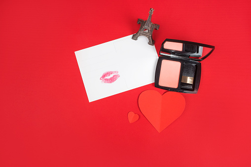 Valentines day flatlay with kiss print postcard, paper heart, blush and eiffel tower figurine isolated on red background.