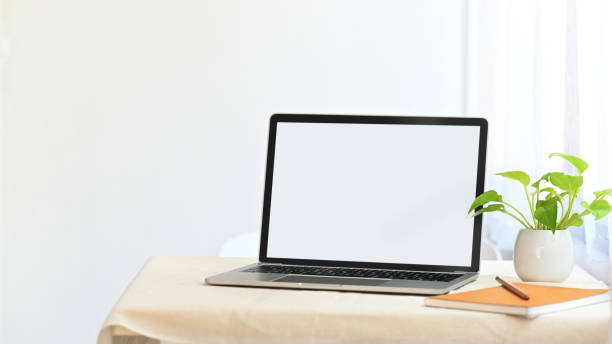 istock Laptop with white blank screen putting together with potted plant, notebook and pencil on the modern table with comfortable living room as background. 1205311145