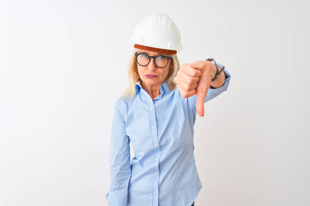istock Middle age architect woman wearing glasses and helmet over isolated white background looking unhappy and angry showing rejection and negative with thumbs down gesture. Bad expression. 1205314667