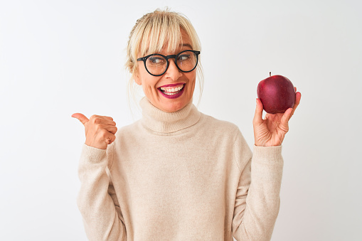 Middle age woman wearing glasses holding apple standing over isolated white background pointing and showing with thumb up to the side with happy face smiling