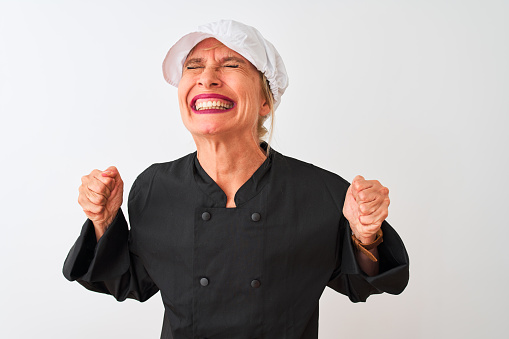 Middle age chef woman wearing uniform and cap standing over isolated white background very happy and excited doing winner gesture with arms raised, smiling and screaming for success. Celebration concept.