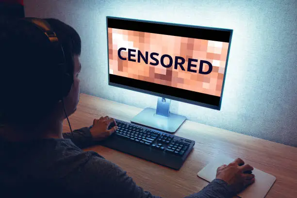 Man in headphones sits in front of monitor with censored content, in dark room