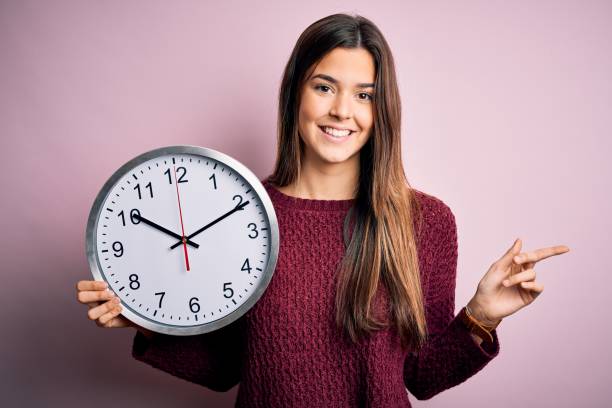 young beautiful girl doing countdown holding big clock over isolated pink background very happy pointing with hand and finger to the side - clock face clock deadline human hand imagens e fotografias de stock