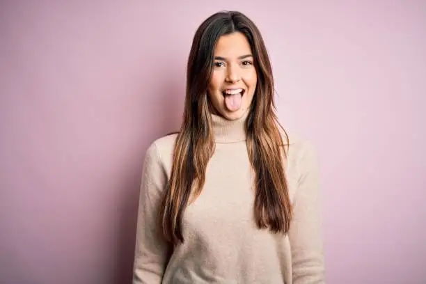 Photo of Young beautiful girl wearing casual turtleneck sweater standing over isolated pink background sticking tongue out happy with funny expression. Emotion concept.