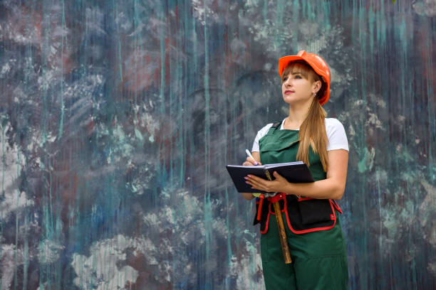 istock Woman in coverall and helmet holding notepad and posing for camera on grey background 1205339233