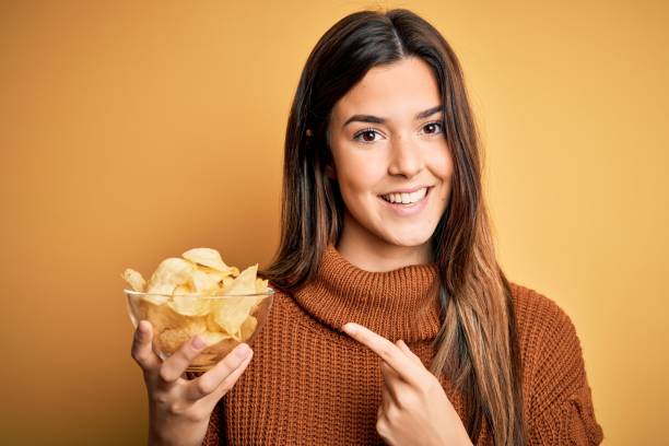 黄色の背景の上に立っているチップスポテトとボウルを保持若い美しい女の子は非常に幸せな手と指で指を指す - potato chip ストックフォトと画像