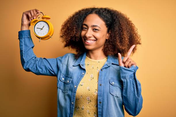 junge afrikanische amerikanische frau mit afro-haar halten vintage wecker über gelben hintergrund überrascht mit einer idee oder frage zeige finger mit glücklichem gesicht, nummer eins - number alarm clock clock hand old fashioned stock-fotos und bilder