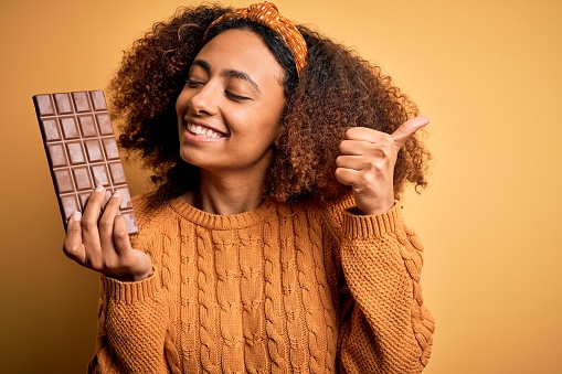 Young african american woman with afro hair holding chocolate bar over yellow background happy with big smile doing ok sign, thumb up with fingers, excellent sign