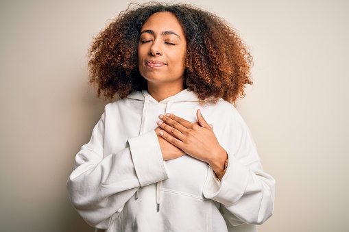 Young african american sportswoman with afro hair wearing sporty sweatshirt smiling with hands on chest with closed eyes and grateful gesture on face. Health concept.