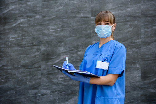 Doctor with notepad. Woman in blue medical coat posing on grey background in mask with stethoscope