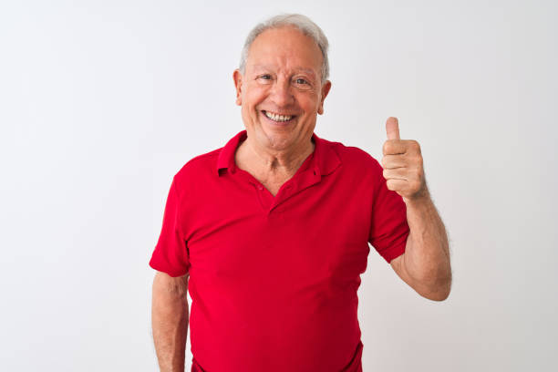 istock Senior grey-haired man wearing red polo standing over isolated white background doing happy thumbs up gesture with hand. Approving expression looking at the camera with showing success. 1205342794