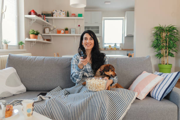 Woman sitting on the couch take some good time with her dog Young beautiful woman watching television with her dog while eating popcorn on the sofa in the living room covering with blanket. Chilling mood plaid couch stock pictures, royalty-free photos & images