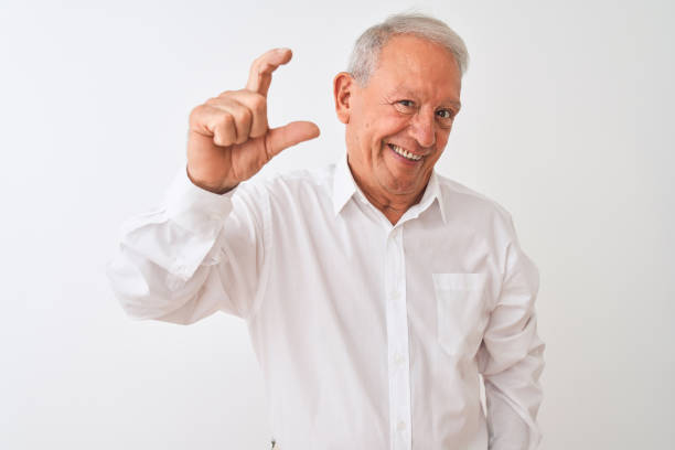 istock Senior grey-haired man wearing elegant shirt standing over isolated white background smiling and confident gesturing with hand doing small size sign with fingers looking and the camera. Measure concept. 1205342943