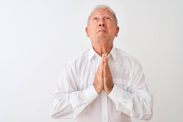 istock Senior grey-haired man wearing elegant shirt standing over isolated white background begging and praying with hands together with hope expression on face very emotional and worried. Asking for forgiveness. Religion concept. 1205342978