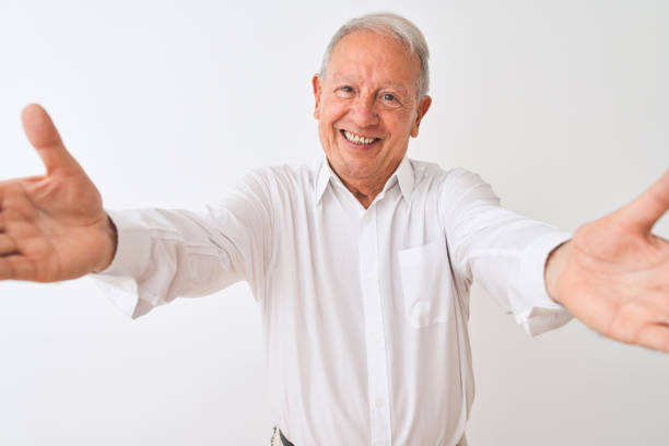 istock Senior grey-haired man wearing elegant shirt standing over isolated white background looking at the camera smiling with open arms for hug. Cheerful expression embracing happiness. 1205343082