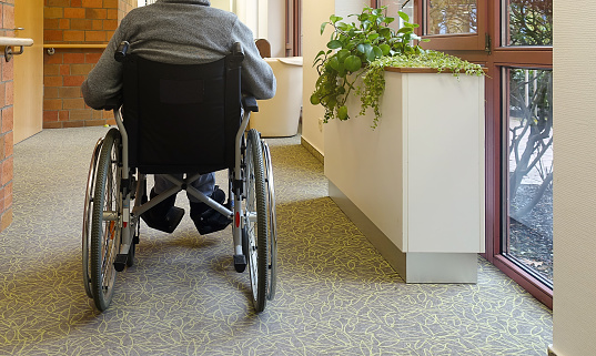 An elderly man, senior, is sitting in a wheelchair in the corridor near the window with his back to the camera. Nursing home.