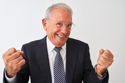 Senior grey-haired businessman wearing suit and glasses over isolated white background very happy and excited doing winner gesture with arms raised, smiling and screaming for success. Celebration concept.