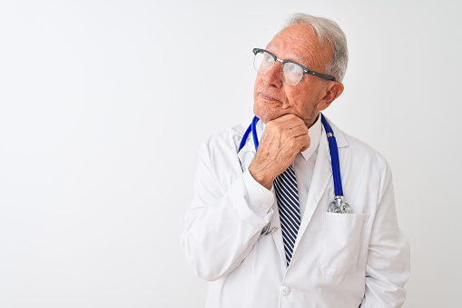 Senior grey-haired doctor man wearing stethoscope standing over isolated white background with hand on chin thinking about question, pensive expression. Smiling with thoughtful face. Doubt concept.