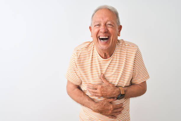 Senior grey-haired man wearing striped t-shirt standing over isolated white background smiling and laughing hard out loud because funny crazy joke with hands on body. Senior grey-haired man wearing striped t-shirt standing over isolated white background smiling and laughing hard out loud because funny crazy joke with hands on body. people laughing hard stock pictures, royalty-free photos & images