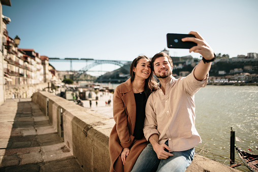 A young couple taking a selfie picture with a modern smartphone. The beautiful river side of Porto, Portugal, appears in the background. Porto is the 2nd city in Portugal and a main touristic destination in the country.