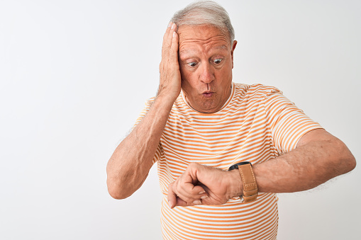 Senior grey-haired man wearing striped t-shirt standing over isolated white background Looking at the watch time worried, afraid of getting late
