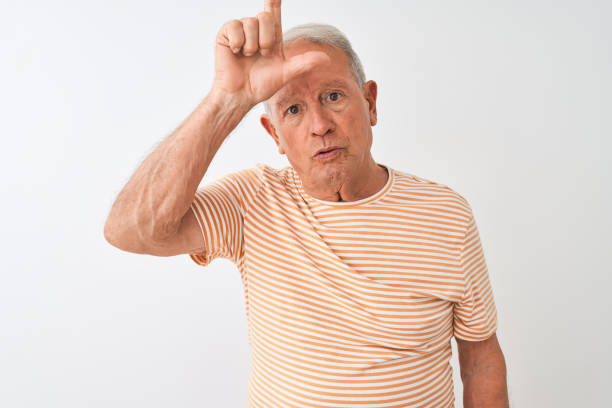 istock Senior grey-haired man wearing striped t-shirt standing over isolated white background making fun of people with fingers on forehead doing loser gesture mocking and insulting. 1205346586