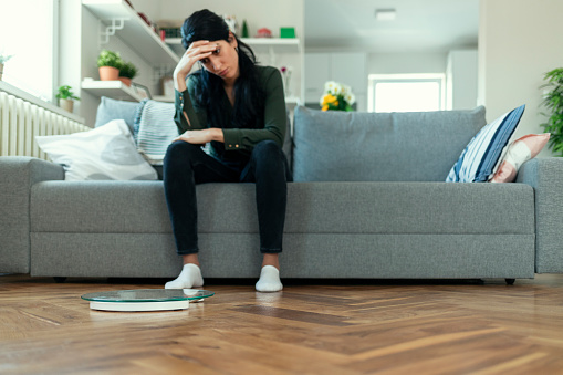 Weighing Scale in Front While Crying Young Woman Sitting on the sofa in the Living Room. Weight Loss Fail Concept. Scale and Depressed, Frustrated and Sad Woman