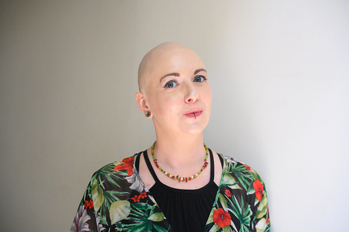 This is a color portrait of a smiling Caucasian Millennial woman in her 30s diagnosed with breast cancer standing by a white wall. She is smiling and looking up with a positive attitude while in the beginning stages of Chemotherapy.