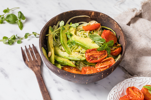 Healthy salad with zoodles zucchini noodles, oven roasted tomatoes and avocado. The salad is served in a recycled environmentally friendly coconut bowl with a wooden fork. Some herbs are sprinkled on the table.