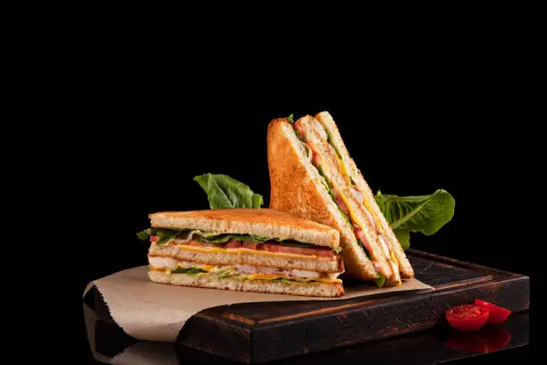 Two halves of a club sandwich on kraft paper. The filling of the sandwich consists of ham, cheese, tomato, chicken breast, sauce and fresh green salad.