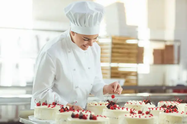 A female confectioner is holding a cake in her hand and in another hand is showing the ok sign in the bakery.