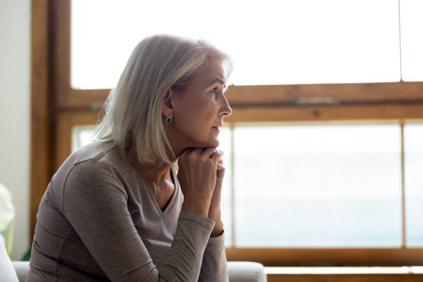 Upset mature woman look in distance feeling sad mourning Side view of sad thoughtful middle-aged mature woman sit on couch at home look in window distance mourning, upset pensive senior female lost in thoughts thinking or pondering over past widow stock pictures, royalty-free photos & images