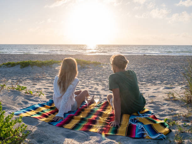 Young couple sharing glass of wine at sunset on beach Two people sharing moment at sunset while relaxing on blanket with a glass of wine outdoors on beach cocoa beach stock pictures, royalty-free photos & images