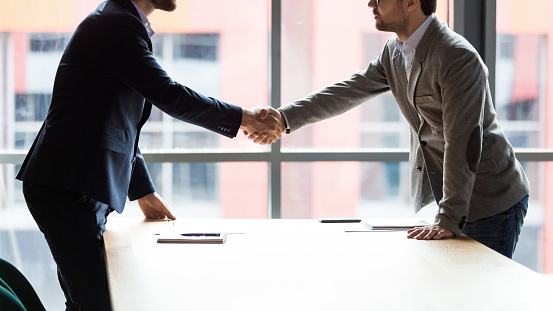 Two businessmen shaking hands at meeting side view, signing partnership contract, legal deal, standing at table in boardroom, business partners greeting, colleagues making agreement at briefing