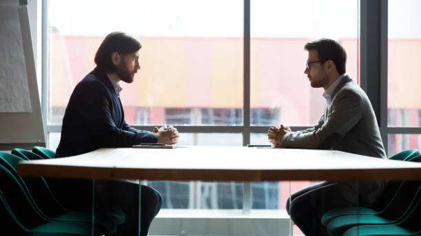 Two businessmen sitting opposite at table, confrontation and negotiation Two serious businessmen sitting opposite at table in boardroom side view, business confrontation and difficult negotiation concept, opponents dialogue debate, competitors looking at each other confrontation stock pictures, royalty-free photos & images