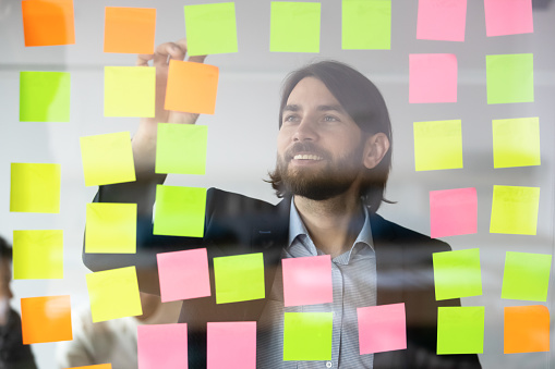 Smiling successful businessman mentor executive reading tasks or new startup ideas on sticky papers, colorful post it notes on glass wall, happy coach planning project on scrum board in office