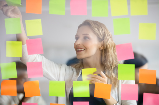 Smiling businesswoman removing sticky papers with finished tasks from scrum board, colorful post it notes, successful happy leader coach planning corporate project at glass wall, adding ideas