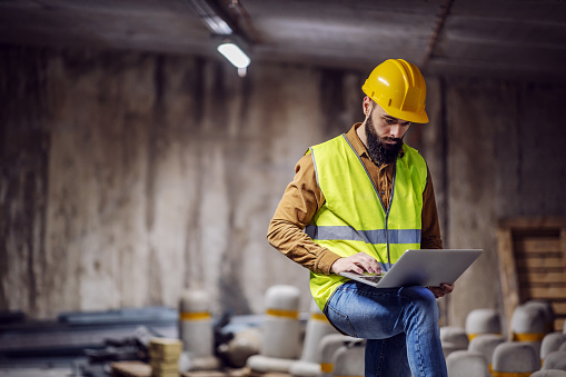 Young attractive focused bearded supervisor in vest with helmet on head standing inside of underground parking lot in construction process and using laptop.